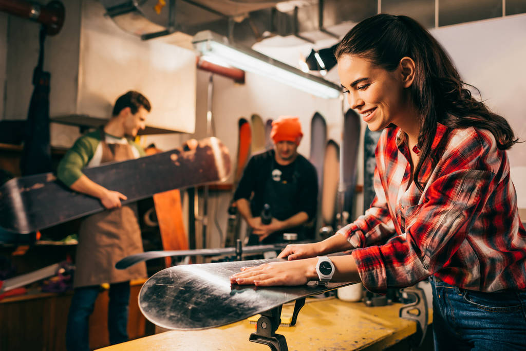Selektiver Fokus lächelnder Arbeiter bei der Reparatur von Snowboards und Kollegen im Hintergrund  - Foto, Bild