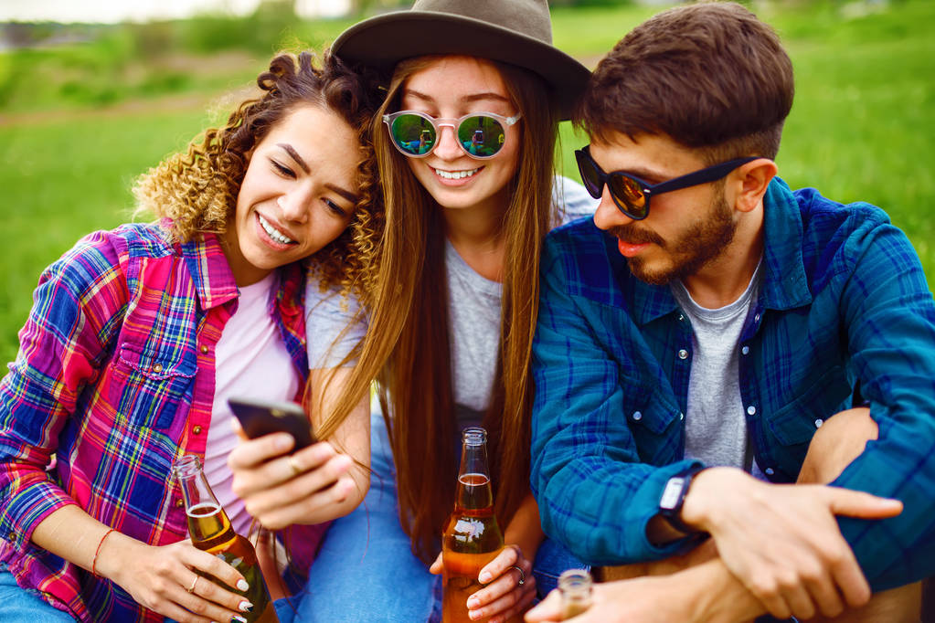 公園で一緒に草の上に座ってビールを飲み、セルフィーを作る幸せな友人。夏の公園でパーティーを楽しむ若者のグループ。休息、楽しさ、夏のコンセプト. - 写真・画像