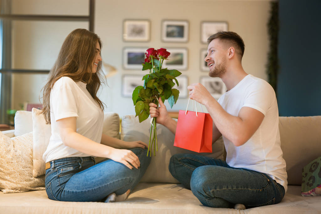 young couple sitting on the sofa, man surprising wife with flowers and gift on saint valentine's day, happy anniversary - Photo, Image