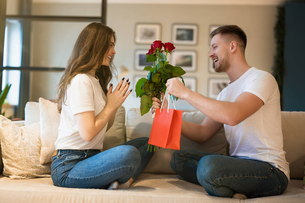 νεαρό υπέροχο ζευγάρι κάθεται στον καναπέ, ο άνθρωπος έκπληξη σύζυγος με λουλούδια και δώρο για την ημέρα του Αγίου Βαλεντίνου, ευτυχισμένη επέτειο - Φωτογραφία, εικόνα