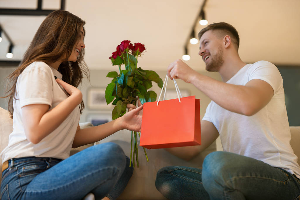 młoda urocza para siedząca na kanapie, mężczyzna zaskakujący żona z różami i prezentem w święte Walentynki, szczęśliwy romantyczny jubileusz - Zdjęcie, obraz