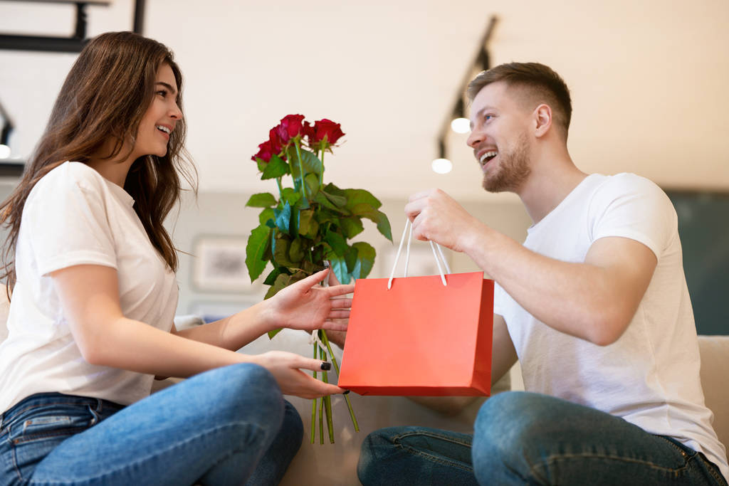 lovely couple sitting on the sofa, handsome husband surprising his beautiful wife with roses and gift on saint valentine's day, happy romantic anniversary - Photo, Image