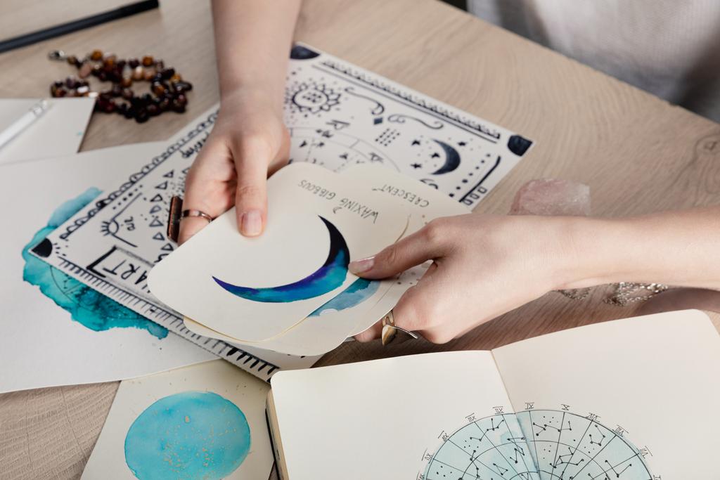 Καλλιεργημένη άποψη του αστρολόγου κρατώντας κάρτες με ακουαρέλα σχέδια από διάγραμμα γέννησης με ζωδιακό κύκλο σημάδι στο τραπέζι - Φωτογραφία, εικόνα