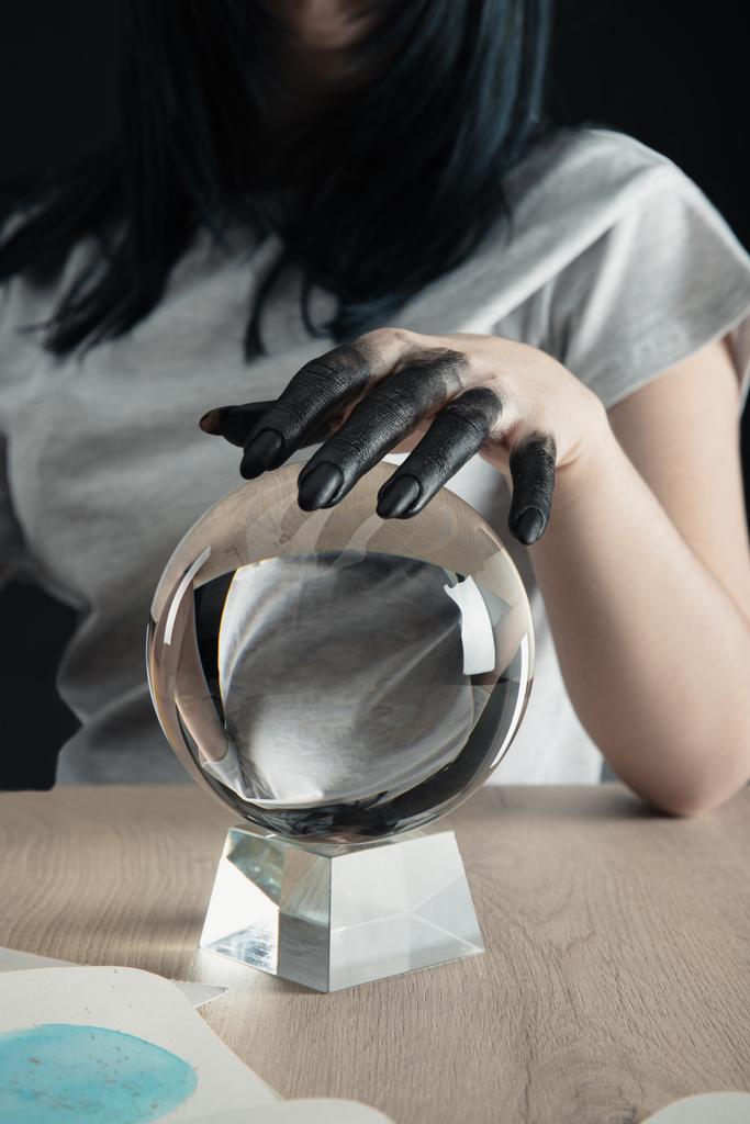 Oříznutý pohled na věštce s černou barvou na ruce držící křišťálovou kouli akvarelovou malbou na stole izolovaném na černém - Fotografie, Obrázek