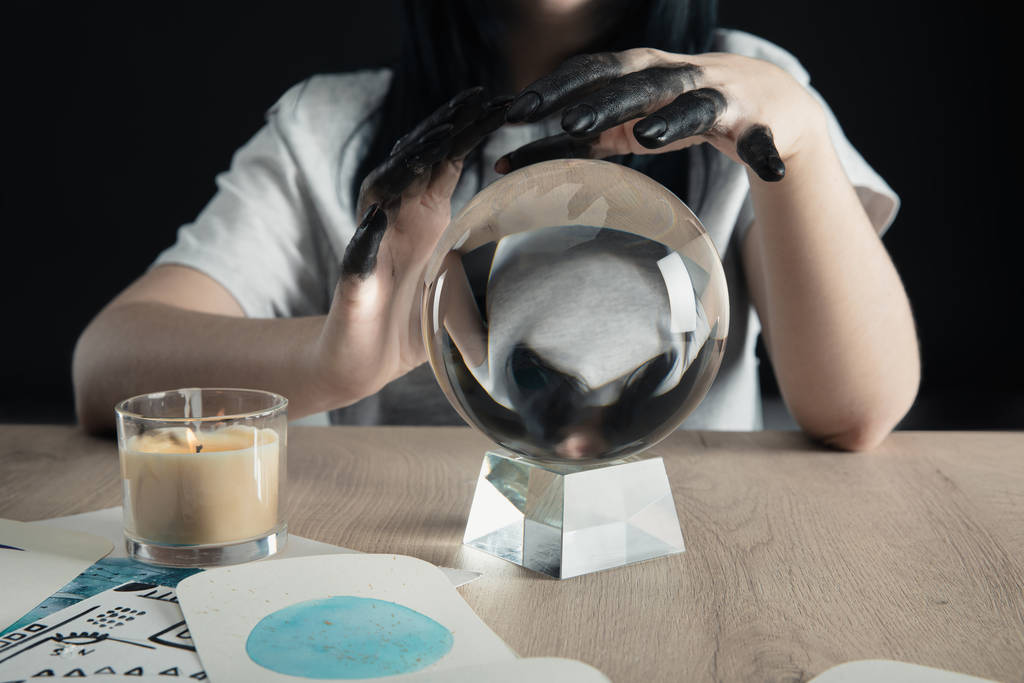 Vista cortada de bruxa com as mãos pintadas de preto, bola de cristal e signos do zodíaco aquarela em cartões na mesa isolada em preto
 - Foto, Imagem