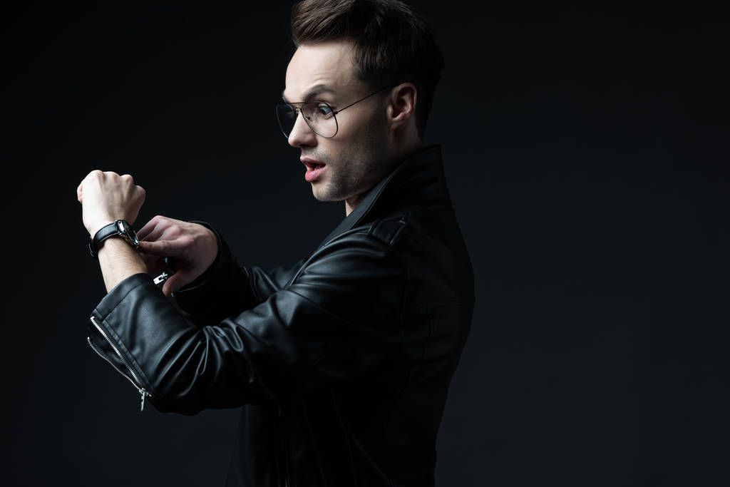 side view of shocked stylish brutal man in biker jacket pointing with finger at wristwatch isolated on black - Photo, Image