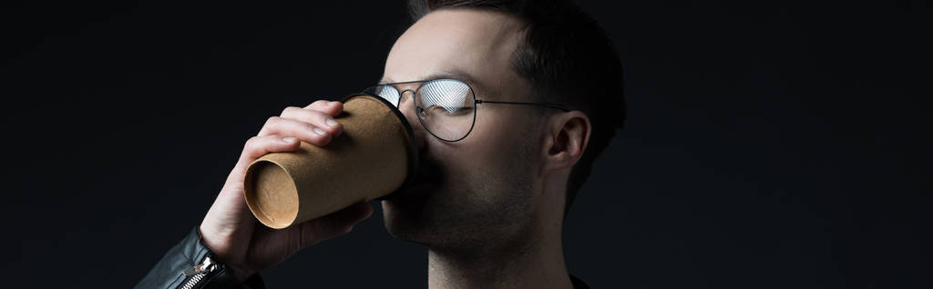 stijlvolle brutale man in bril drinken koffie uit papieren beker geïsoleerd op zwart, panoramisch schot - Foto, afbeelding