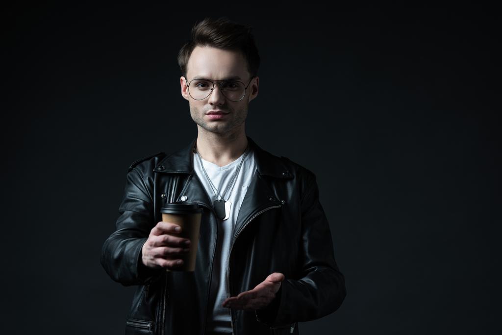 stylish brutal man in biker presenting coffee in paper cup isolated on black - Photo, Image