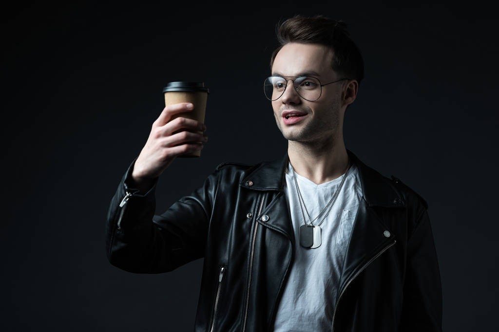 excited stylish brutal man in biker jacket looking at coffee to go isolated on black - Photo, Image