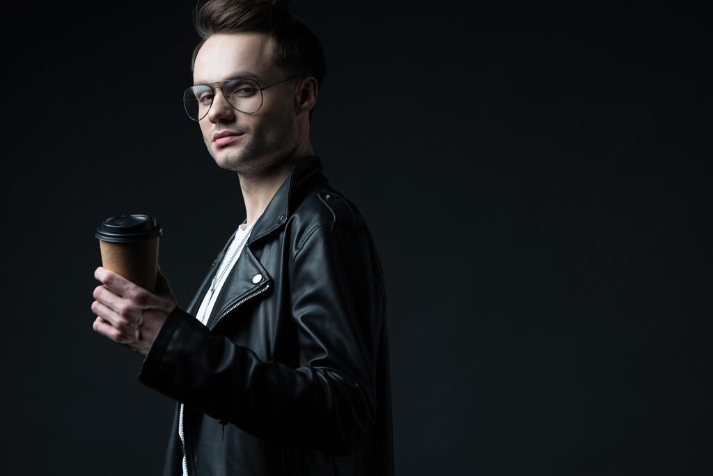 side view of stylish brutal man in biker jacket with coffee to go isolated on black - Photo, Image