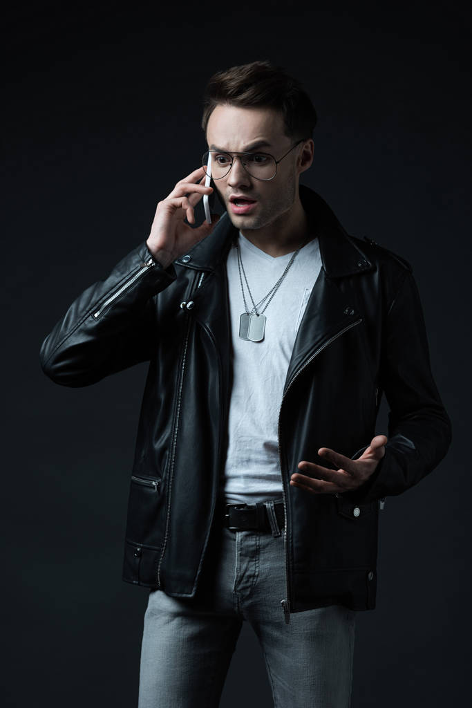 shocked tense stylish man in leather jacket talking on smartphone isolated on black - Photo, Image