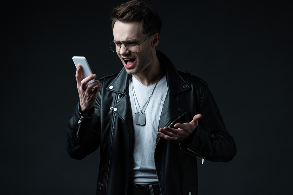 angry stylish man in leather jacket yelling at smartphone isolated on black - Photo, Image