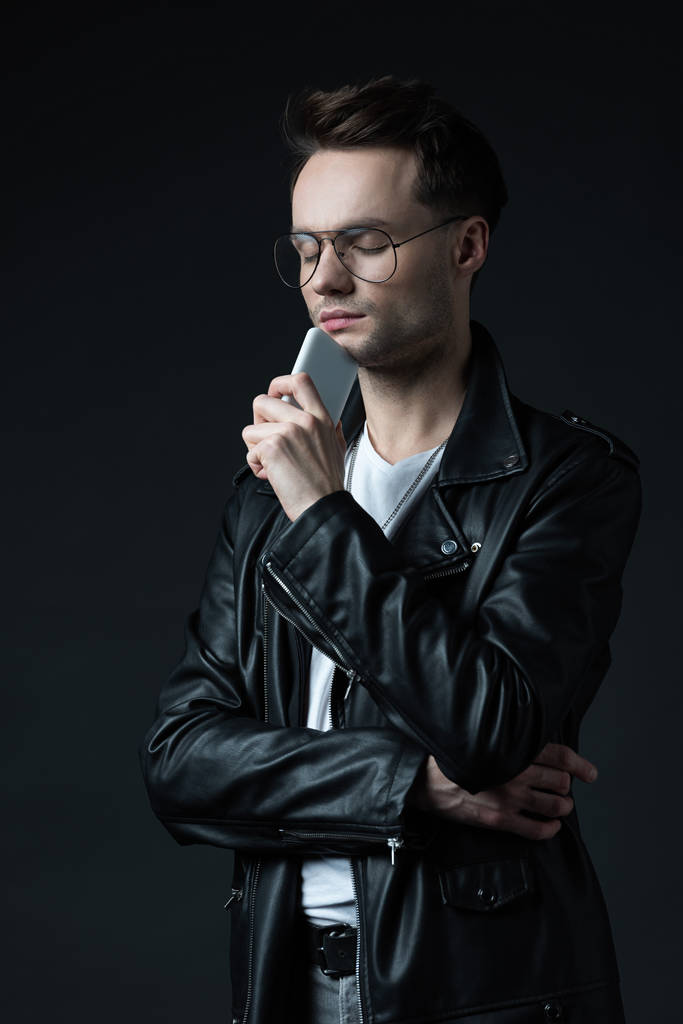 homem elegante pensativo em jaqueta de couro com smartphone e olhos fechados isolados em preto
 - Foto, Imagem