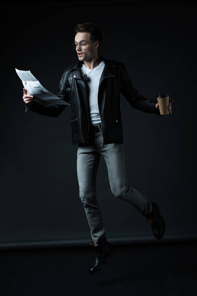 scioccato uomo brutale elegante in giacca da motociclista levitante con tazza di carta e giornale isolato su nero
 - Foto, immagini