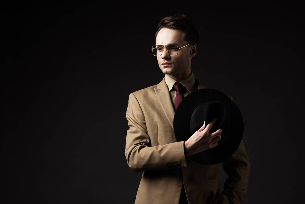 hombre elegante pensativo en traje beige y gafas con sombrero aislado en negro
 - Foto, imagen