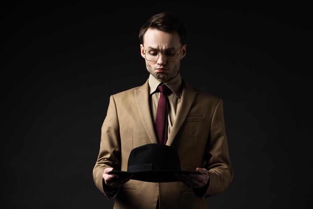 elegantní muž v béžovém obleku a brýlích držící klobouk a šklebící se izolovaně na černé - Fotografie, Obrázek