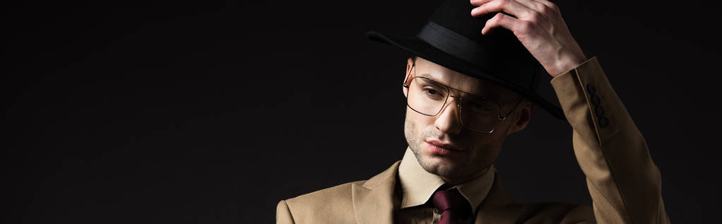 elegant man in beige suit and eyeglasses putting on hat isolated on black, panoramic shot - Photo, Image