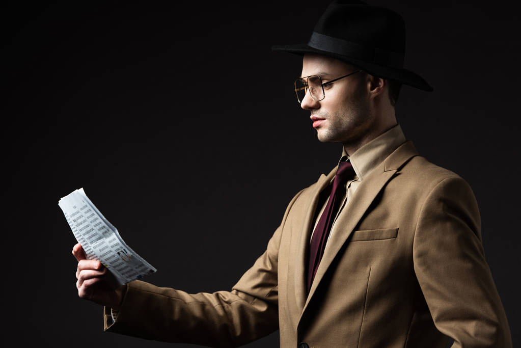 Seitenansicht eines eleganten Mannes in beigem Anzug, Hut und Brille beim Zeitungslesen isoliert auf schwarz - Foto, Bild