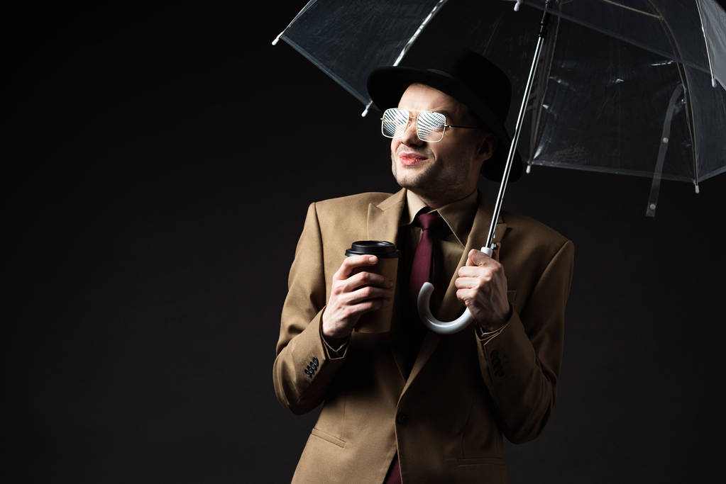 confus homme élégant en costume beige, chapeau et lunettes tenant parapluie et café pour aller isolé sur noir
 - Photo, image