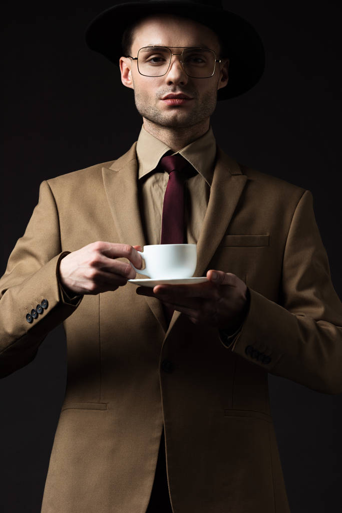 elegante man in beige pak met koffiekop en schotel geïsoleerd op zwart - Foto, afbeelding