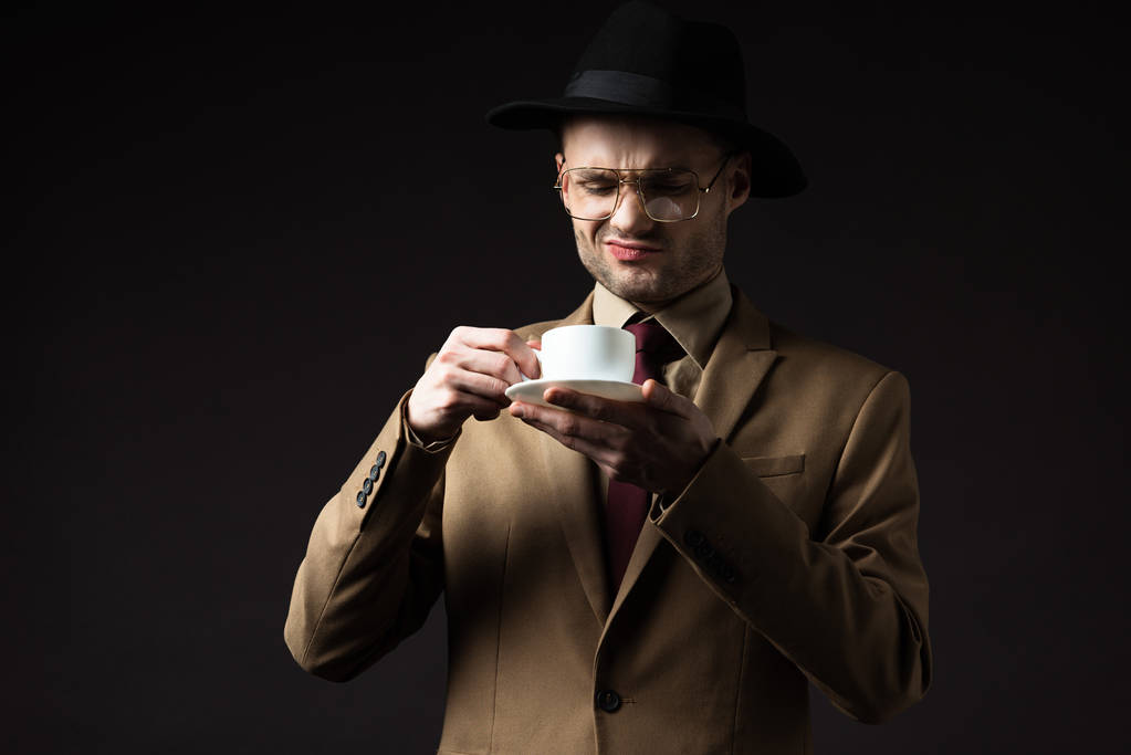 sceptyczny elegancki mężczyzna w beżowym garniturze, kapeluszu i okularach trzymający filiżankę kawy i spodek odizolowany na czarno - Zdjęcie, obraz