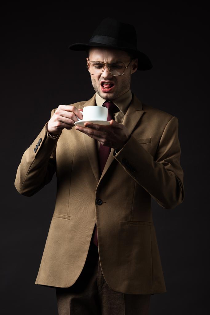 displeased elegant man in beige suit, hat and eyeglasses holding coffee cup and saucer isolated on black - Photo, Image