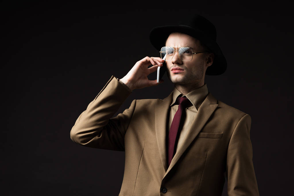 ernstige elegante man in beige pak, hoed en bril praten op smartphone geïsoleerd op zwart - Foto, afbeelding