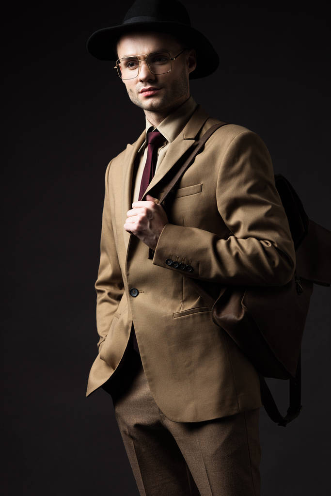 homme élégant sérieux en costume beige, chapeau et lunettes tenant sac à dos en cuir brun isolé sur noir
 - Photo, image