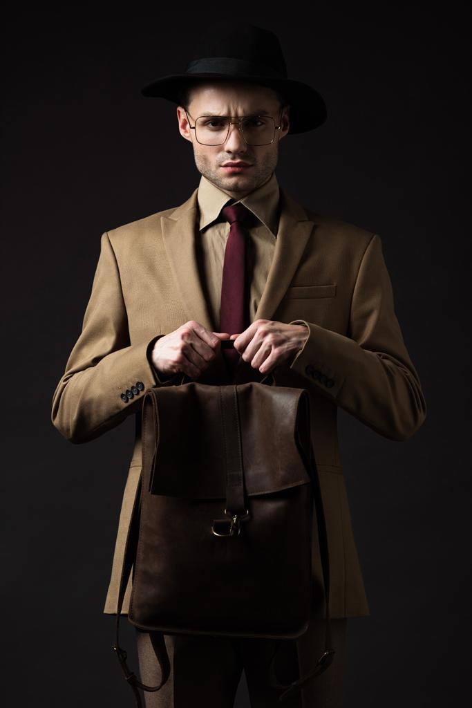 napięty elegancki mężczyzna w beżowym garniturze, kapeluszu i okularach trzymający brązowy skórzany plecak odizolowany na czarno - Zdjęcie, obraz