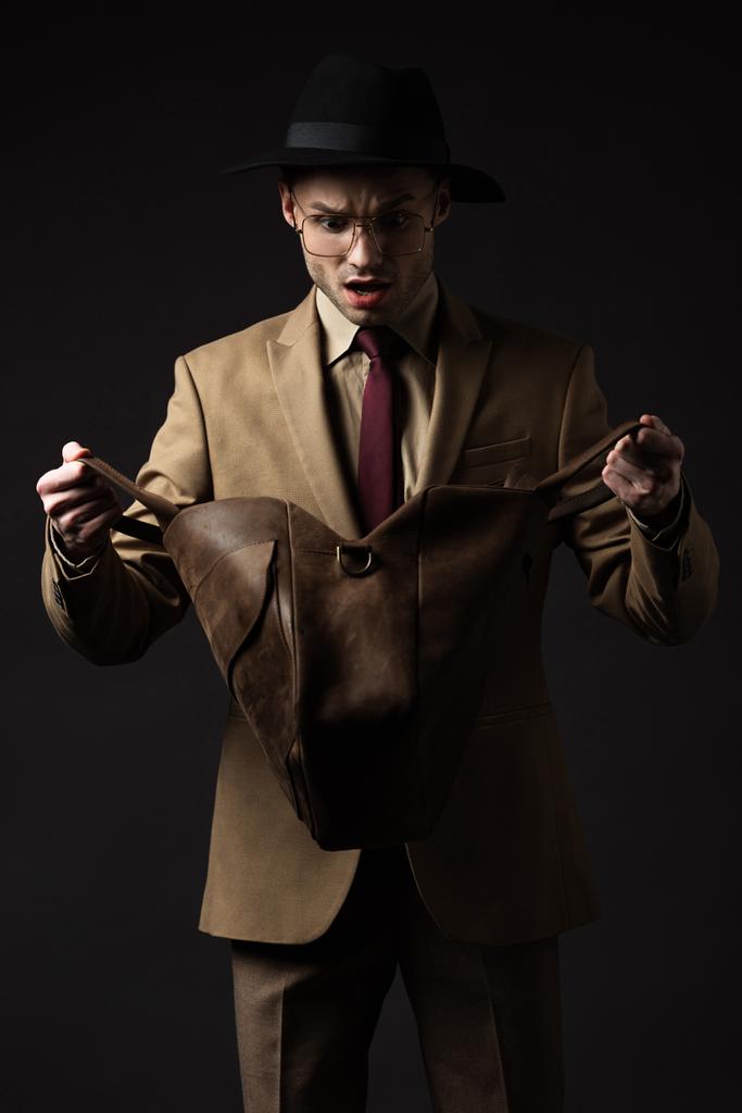 homme élégant choqué en costume beige, chapeau et lunettes à la recherche dans un sac en cuir brun isolé sur noir
 - Photo, image