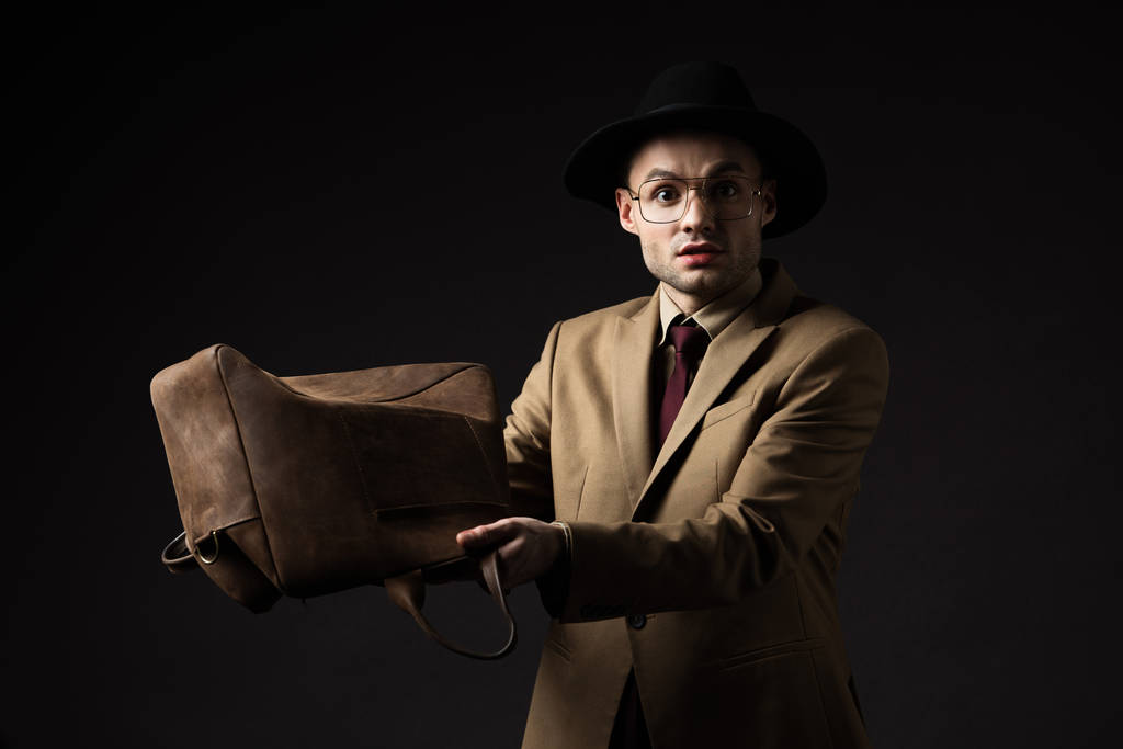 sorprendido hombre elegante en traje beige, sombrero y gafas sacuden bolsa de cuero marrón aislado en negro
 - Foto, Imagen