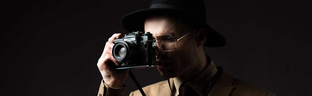 κομψός άνδρας σε μπεζ κοστούμι, καπέλο και γυαλιά που τραβά φωτογραφία σε κάμερα που απομονώνεται σε μαύρο, πανοραμικό πλάνο - Φωτογραφία, εικόνα