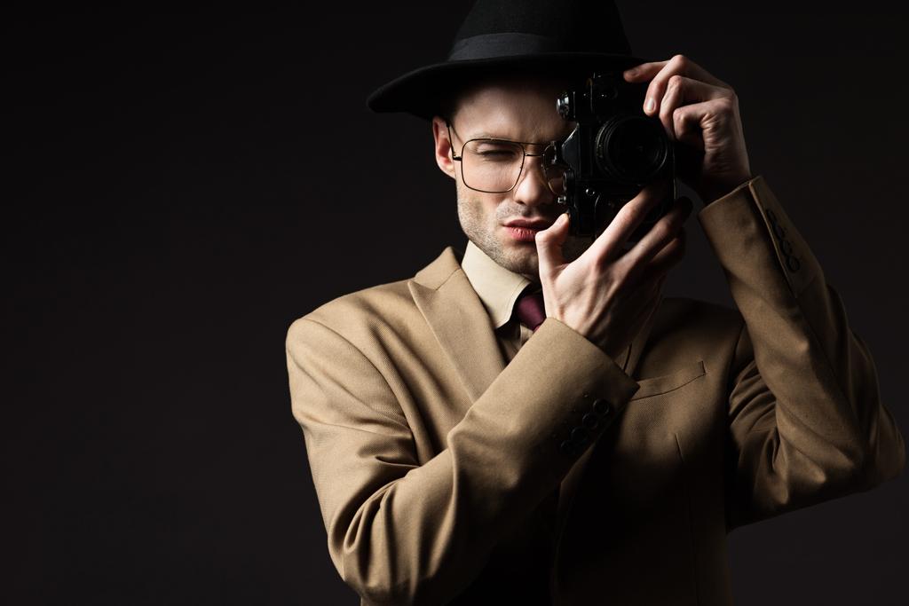 elegant man in beige suit, hat and eyeglasses taking picture on film camera isolated on black - Photo, Image