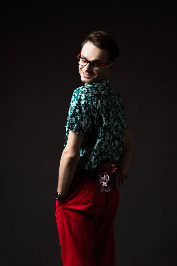 uomo alla moda sorridente in occhiali in camicia colorata blu e pantaloni rossi tenendo bouquet floreale dietro la schiena isolato su nero
 - Foto, immagini