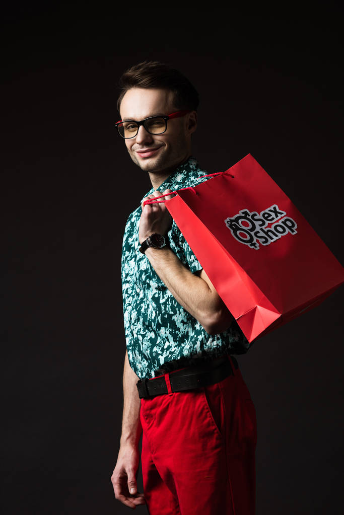 sonriente hombre de moda en gafas de vista en camisa de colores azules y pantalones rojos sosteniendo bolsa de compras roja de sex shop aislado en negro
 - Foto, Imagen
