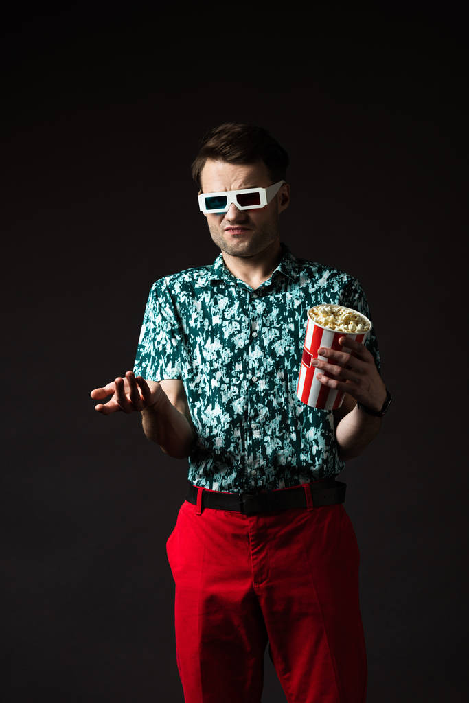 zdezorientowany modny mężczyzna w okularach 3d w niebieskiej koszuli i czerwonych spodniach trzymając popcorn odizolowany na czarno - Zdjęcie, obraz