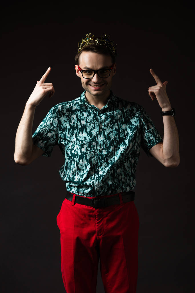 χαμογελαστός μοντέρνος άντρας με γυαλιά που δείχνει με τα δάχτυλα στο στέμμα απομονωμένα στο μαύρο - Φωτογραφία, εικόνα