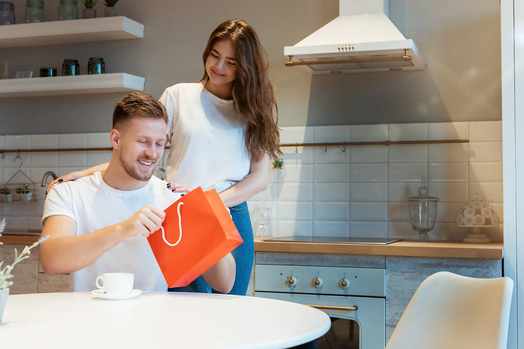 šťastná krásná brunetka žena pozdravuje svého překvapeného manžela s dárkem v červené tašce na svatého Valentýna při snídani v kuchyni, šťastné romantické výročí - Fotografie, Obrázek