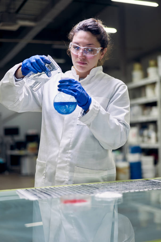 Молодая лаборантка в очках и белом халате с фляжкой с голубой жидкостью в руках проводит эксперименты
 - Фото, изображение