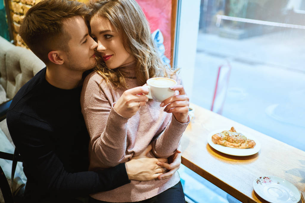 Kafenin penceresinde oturan, kucaklaşan, öpüşen ve kahve içen mutlu romantik çift. Sevgili, sıcak kafede kız arkadaşına şefkatle sarılıyor.. - Fotoğraf, Görsel