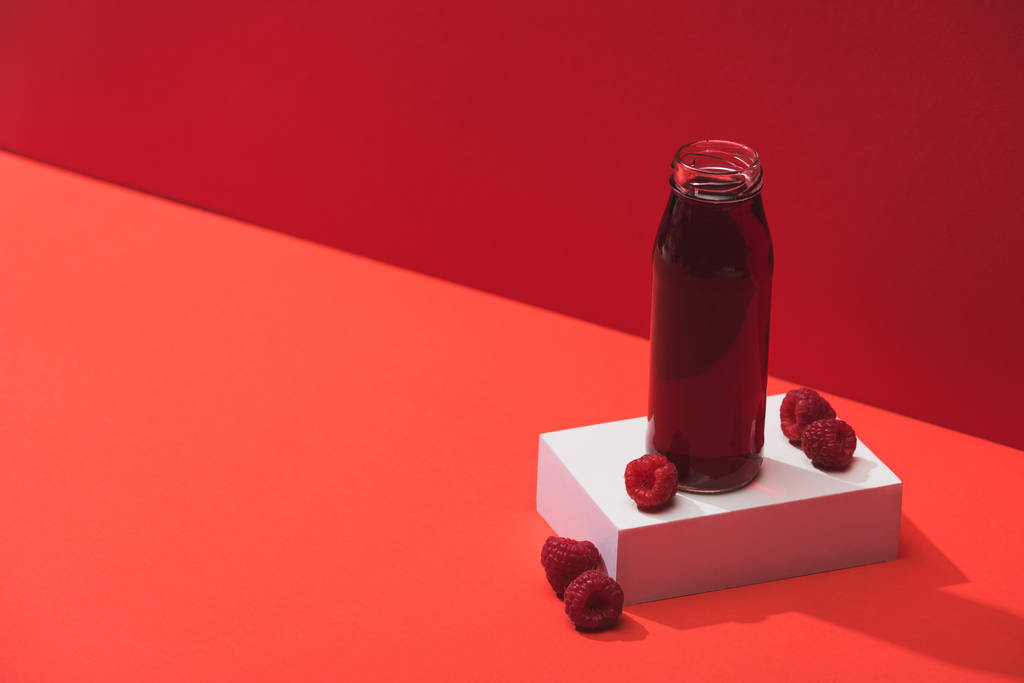 zumo de bayas frescas en botella de vidrio cerca de frambuesas maduras en cubo sobre fondo rojo
 - Foto, Imagen