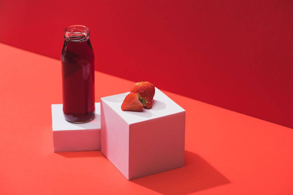 φρέσκος χυμός μούρου σε γυάλινο μπουκάλι κοντά ώριμη φράουλα σε κύβους σε κόκκινο φόντο - Φωτογραφία, εικόνα