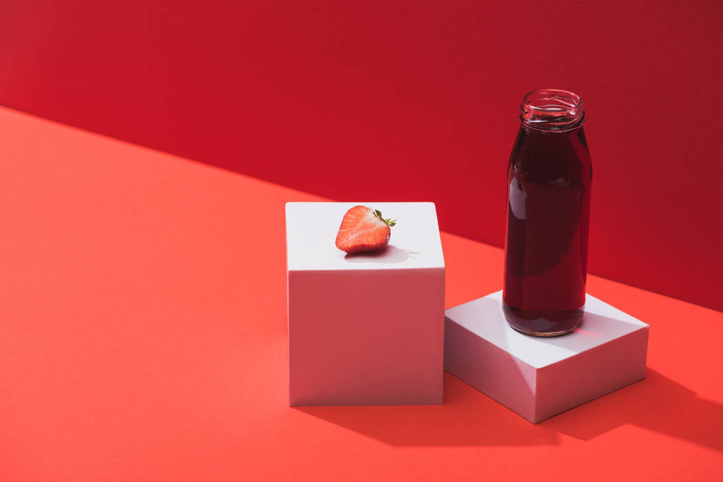 świeży sok jagodowy w szklanej butelce w pobliżu dojrzałej truskawki na kostkach na czerwonym tle - Zdjęcie, obraz