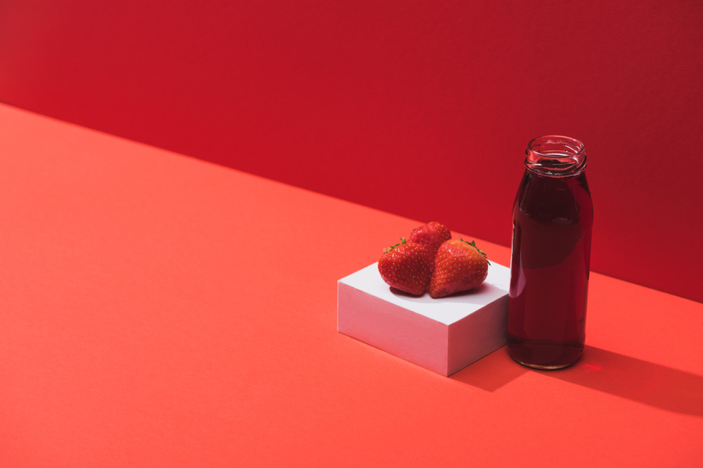 Свежий ягодный сок в стеклянной бутылке возле спелой клубники на кубе на красном фоне
 - Фото, изображение