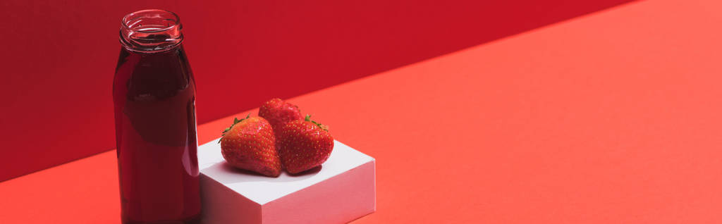φρέσκο χυμό μούρου σε γυάλινο μπουκάλι κοντά ώριμες φράουλες σε κύβο σε κόκκινο φόντο, πανοραμική λήψη - Φωτογραφία, εικόνα
