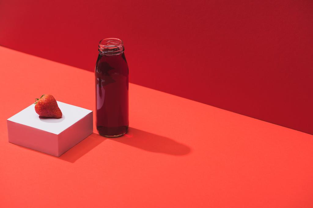 φρέσκος χυμός μούρου σε γυάλινο μπουκάλι κοντά ώριμη φράουλα σε κύβο σε κόκκινο φόντο - Φωτογραφία, εικόνα