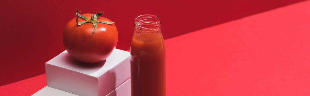 fresh vegetable juice in glass bottle near ripe tomato on stand on red background, panoramic shot - Photo, Image