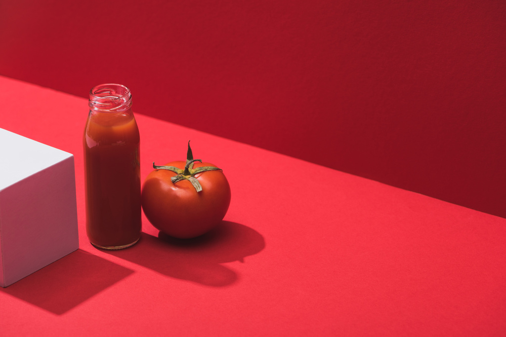 Свежий овощной сок в стеклянной бутылке возле спелых помидоров и куба на красном фоне
 - Фото, изображение