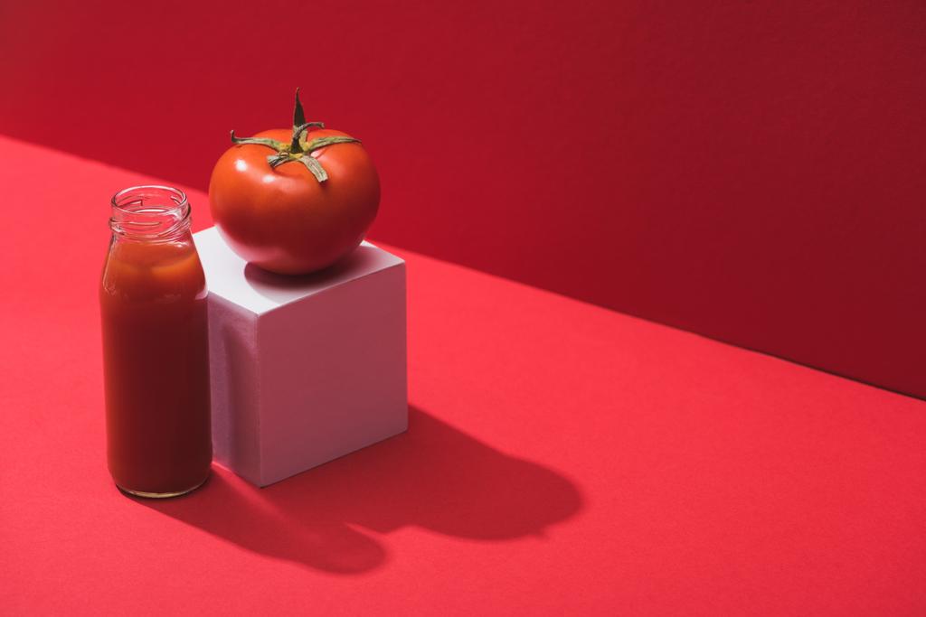 φρέσκος χυμός λαχανικών σε γυάλινο μπουκάλι και ώριμη ντομάτα σε κύβο σε κόκκινο φόντο - Φωτογραφία, εικόνα