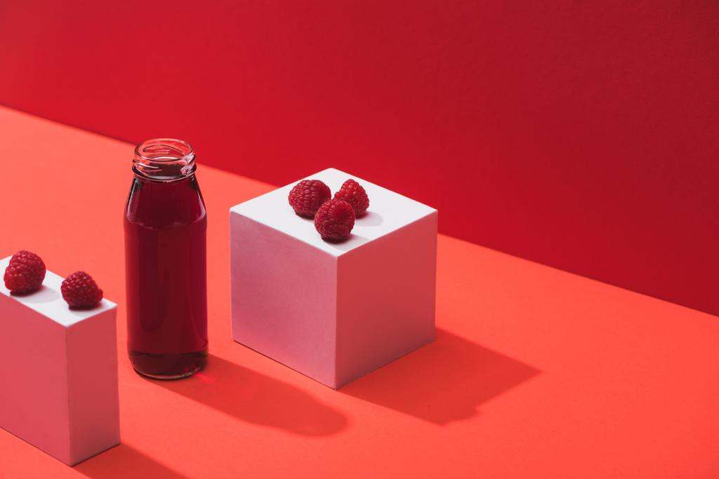 fresh berry juice in glass bottle near ripe raspberries on cubes on red background - Photo, Image